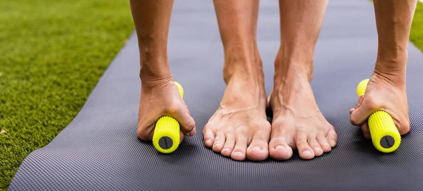Person uses a bright yellow massage roller on a black exercise mat, feet flat on the mat in a stretch setup