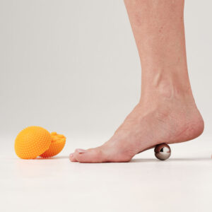 Barefoot foot resting on a small metal massage ball with bright orange spiky massage balls nearby on a white surface.
